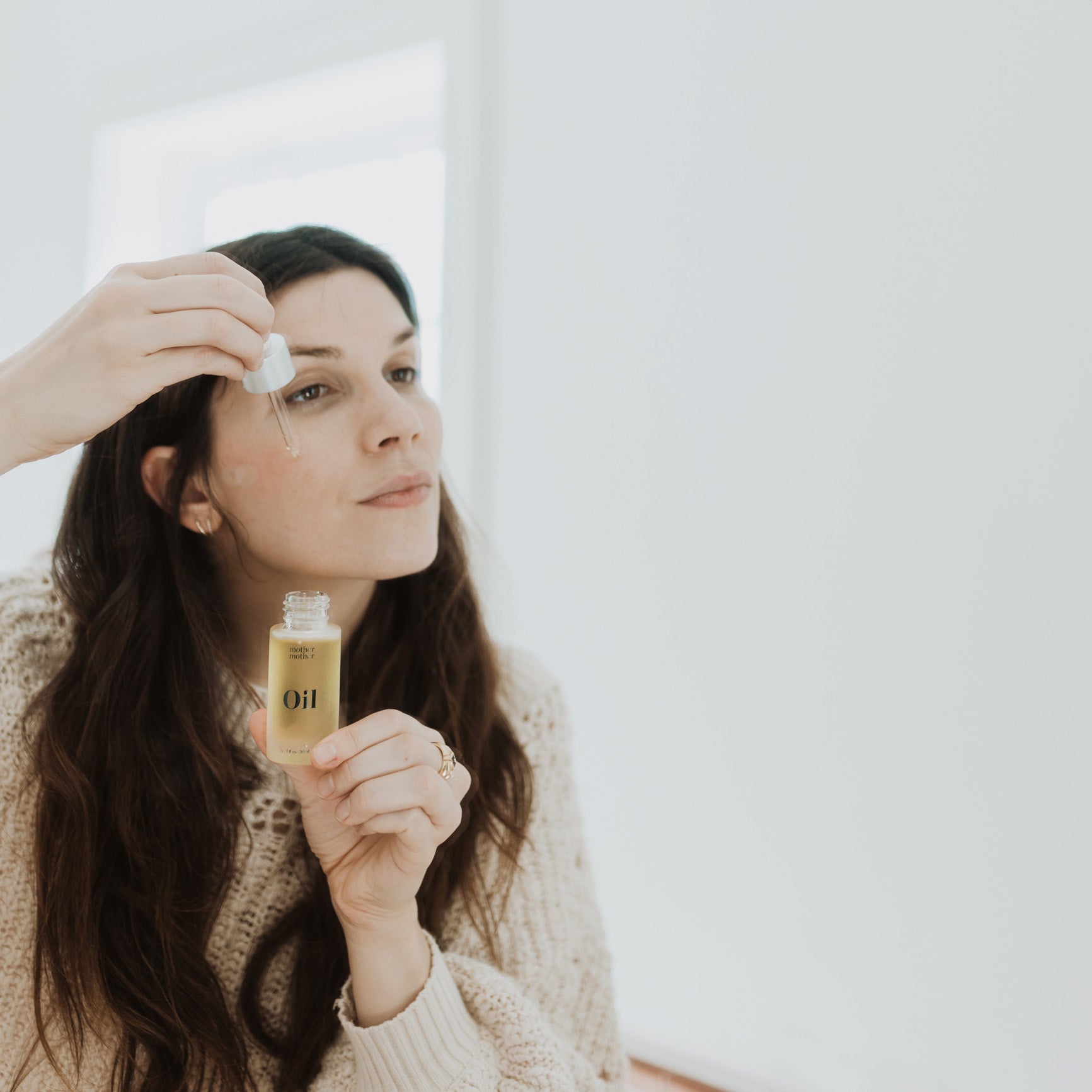 woman with brown hair and beige sweater putting mother mother oil on her face